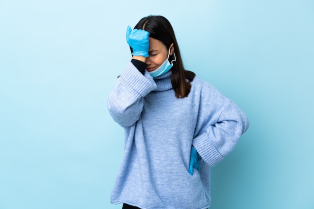 Junge brünette Mischrassenfrau, die mit einer Maske und Handschuhen über blauer Wand mit Kopfschmerzen schützt