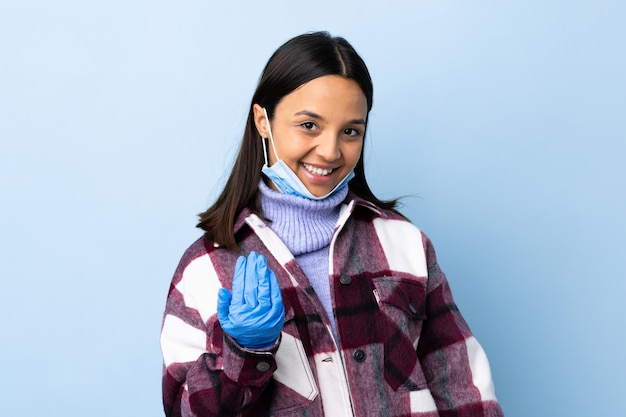 Junge brünette Mischrassenfrau, die mit einer Maske und Handschuhen über blauer Wand schützt, die einlädt, mit Hand zu kommen.