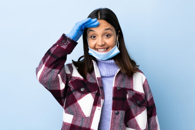 Junge brünette Mischrassenfrau, die mit einer Maske und Handschuhen über blauer Wand schützt, die mit Hand mit glücklichem Ausdruck salutiert