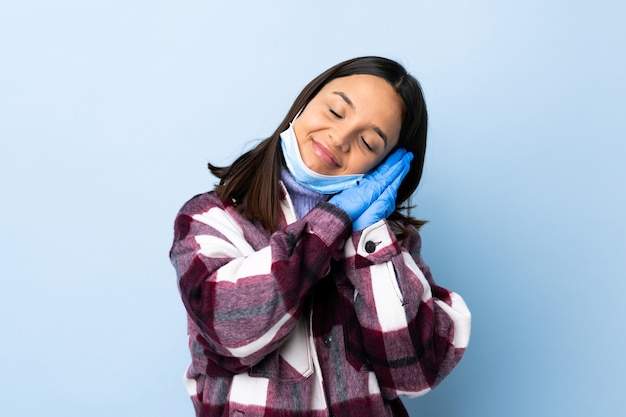 Junge brünette Mischrassenfrau, die mit einer Maske und Handschuhen über blauer Wand schützt, die Schlafgeste im entzückenden Ausdruck macht