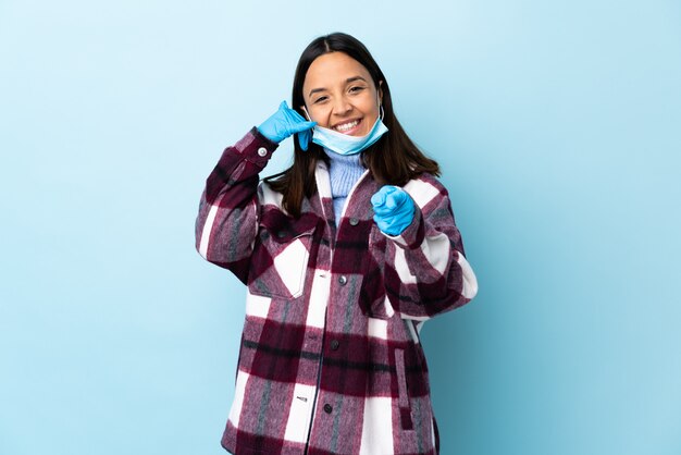 Junge brünette Mischrassenfrau, die mit einer Maske und Handschuhen über der blauen Wand schützt, die Telefongeste macht und nach vorne zeigt