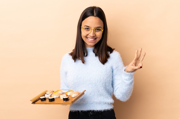 Junge brünette Mischrassenfrau, die Sushi über isolierte Wand glücklich hält und drei mit den Fingern zählt