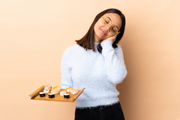 Junge brünette Mischrassenfrau, die Sushi über isolierter Wand hält, die Schlafgeste in dorable Ausdruck macht