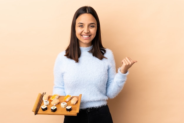 Junge brünette Mischrassenfrau, die Sushi über Wand hält, die zur Seite zeigt, um ein Produkt zu präsentieren