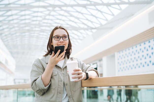 Junge brünette Teenager-Studentin in Brille mit Handy sendete Sprachnachricht mit Kaffeetasse aus Papier im Einkaufszentrum an öffentlichen Orten