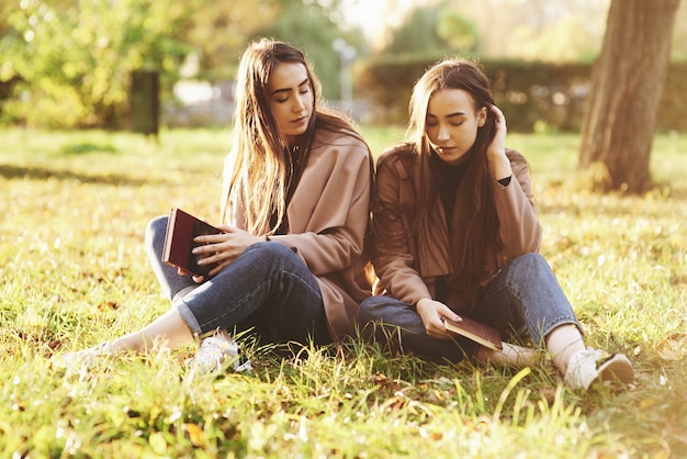 Junge brünette Zwillingsschwestern sitzen nahe beieinander, die Augen auf dem Gras geschlossen, die Beine leicht in die Knie gebeugt und gekreuzt, braune Bücher haltend, lässigen Mantel im Herbstpark auf Hintergrund tragend.