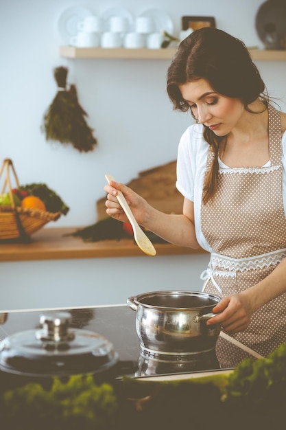Junge Brunettefrau, die Suppe in der Küche kocht. Hausfrau, die hölzernen Löffel in ihrer Hand hält. Lebensmittel- und Gesundheitskonzept.