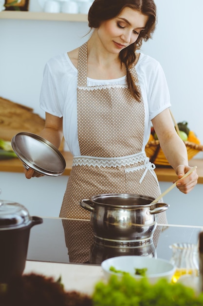 Junge Brunettefrau, die Suppe in der Küche kocht. Hausfrau, die hölzernen Löffel in ihrer Hand hält. Lebensmittel- und Gesundheitskonzept.