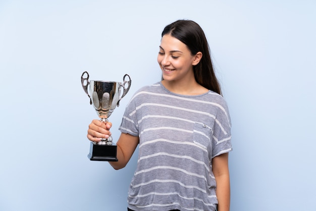 Junge Brunettefrau über der lokalisierten blauen Wand, die eine Trophäe hält