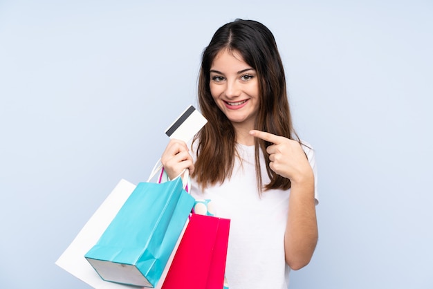 Junge Brunettefrau über der lokalisierten blauen Wand, die Einkaufstaschen und eine Kreditkarte hält