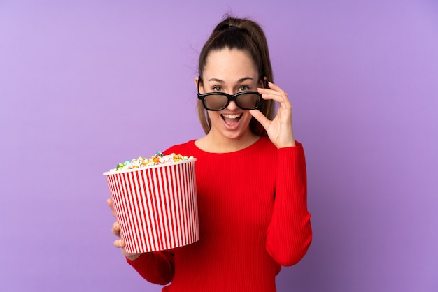 Junge Brunettefrau über lokalisierter purpurroter Wand überrascht mit Gläsern 3d und einem großen Eimer Popcorn halten