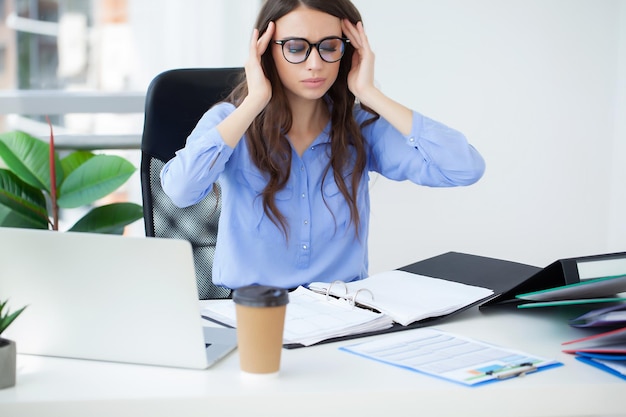 Junge Büroangestellte, die wegen Schmerzen im Büro am Schreibtisch sitzen und den Kopf halten