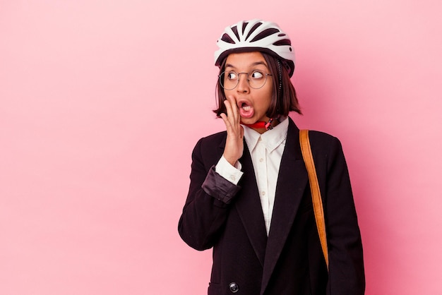 Junge Business-Mixed-Race-Frau, die einen Fahrradhelm auf rosafarbenem Hintergrund trägt, sagt eine geheime heiße Bremsnachricht und schaut beiseite