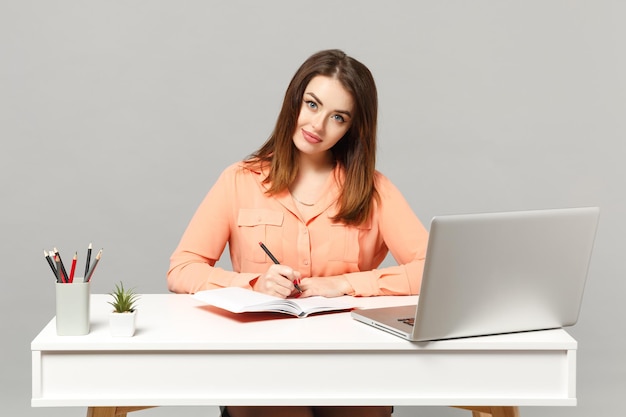 Junge charmante Frau in pastellfarbener Freizeitkleidung schreibt Notizen, sitzt und arbeitet am weißen Schreibtisch mit PC-Laptop isoliert auf grauem Hintergrund. Leistung Business-Karriere-Lifestyle-Konzept. Mock-up-Kopienbereich.