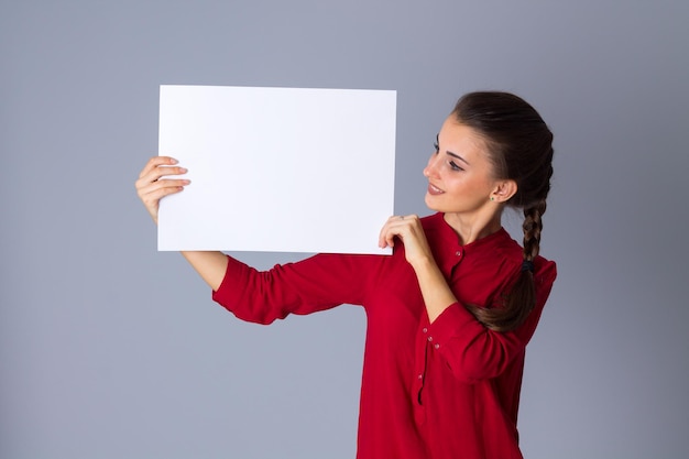 Junge charmante Frau in roter Bluse mit weißem Blatt Papier auf grauem Hintergrund im Studio