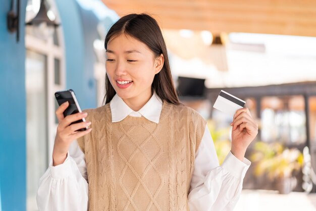 Junge Chinesin beim Einkaufen im Freien mit dem Handy mit Kreditkarte