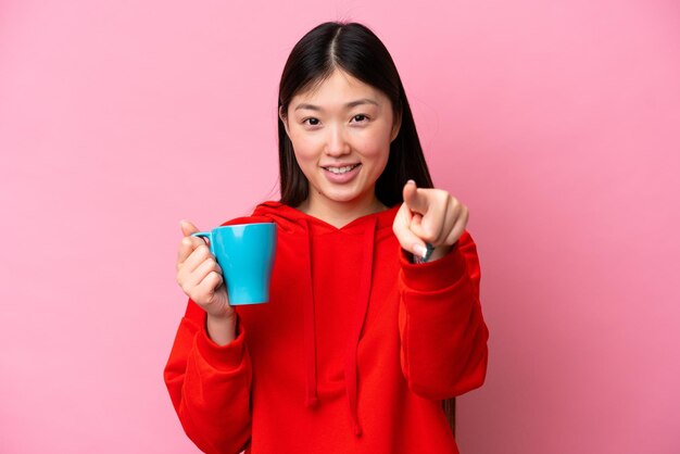 Junge Chinesin, die eine Tasse Kaffee isoliert auf rosa Hintergrund hält, zeigt mit einem selbstbewussten Ausdruck mit dem Finger auf Sie