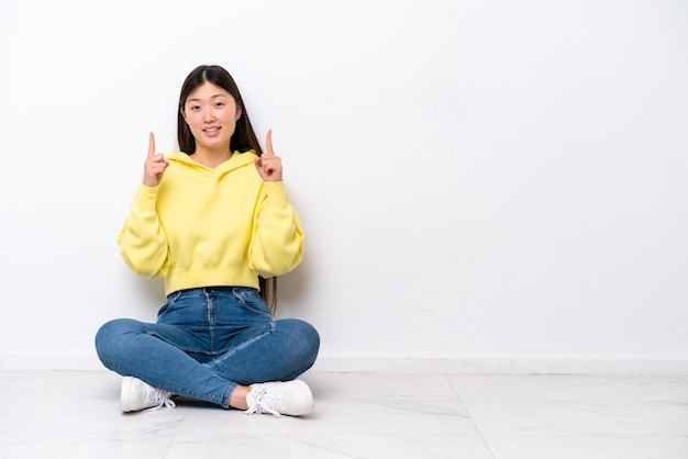 Junge Chinesin, die isoliert auf dem Boden auf einer weißen Wand sitzt und eine großartige Idee zeigt