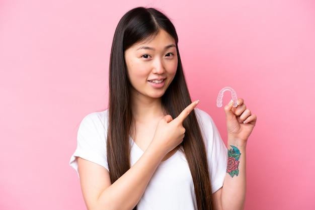 Junge Chinesin, die unsichtbare Zahnspangen isoliert auf rosa Hintergrund hält und darauf zeigt