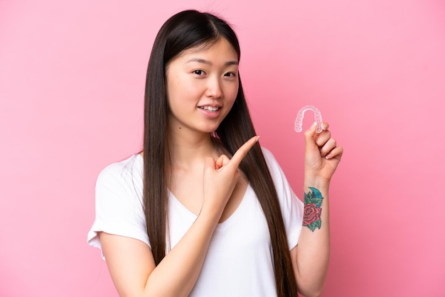 Junge Chinesin, die unsichtbare Zahnspangen isoliert auf rosa Hintergrund hält und darauf zeigt