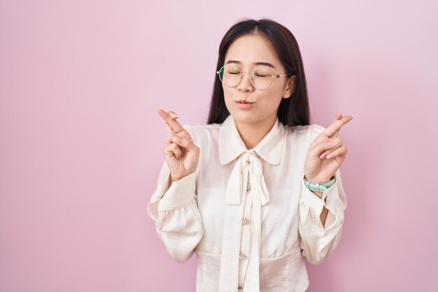 Junge Chinesin, die vor rosa Hintergrund steht und mit gekreuzten Fingern lächelnd und hoffnungsvoll mit geschlossenen Augen gestikuliert. Glück und abergläubisches Konzept.