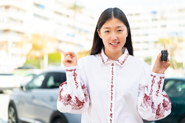 Junge Chinesin hält Autoschlüssel im Freien überrascht und zeigt mit dem Finger zur Seite