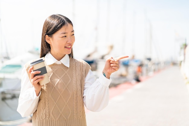 Junge Chinesin hält draußen einen Kaffee zum Mitnehmen in der Hand und zeigt zur Seite, um ein Produkt zu präsentieren