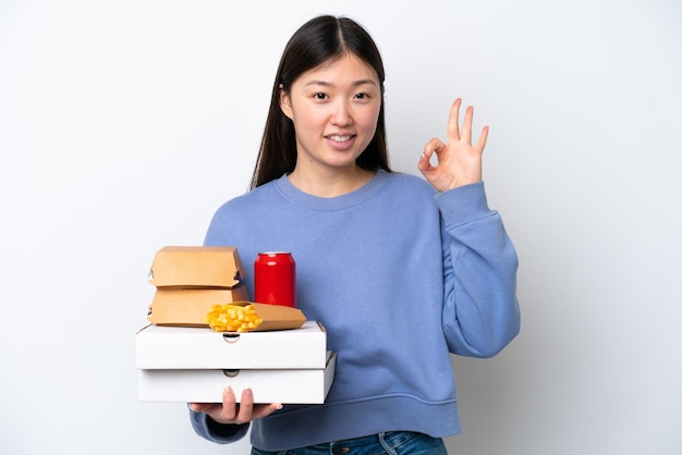 Junge Chinesin hält Fast Food isoliert auf weißem Hintergrund und zeigt Ok-Zeichen mit den Fingern