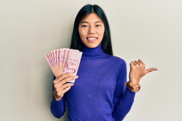 Junge Chinesin hält thailändische Baht-Banknoten in der Hand, zeigt mit dem Daumen zur Seite und lächelt glücklich mit offenem Mund