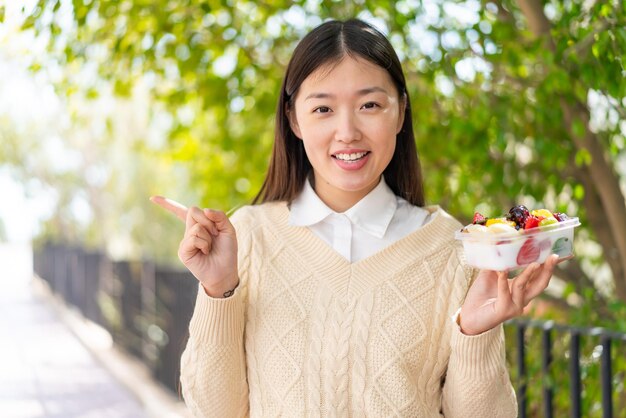 Junge Chinesin hält überrascht eine Obstschale im Freien und zeigt mit dem Finger zur Seite