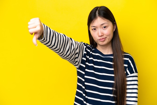 Junge Chinesin isoliert auf gelbem Hintergrund, die Daumen nach unten mit negativem Ausdruck zeigt