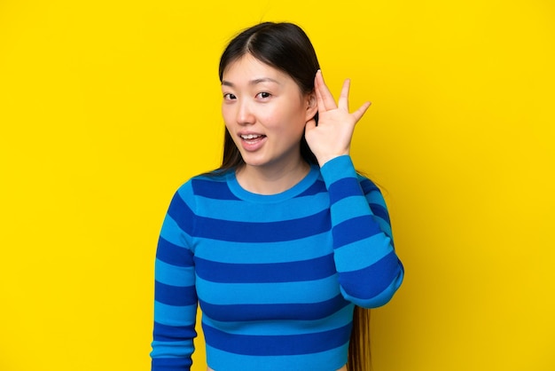 Junge Chinesin isoliert auf gelbem Hintergrund, die etwas hört, indem sie die Hand auf das Ohr legt