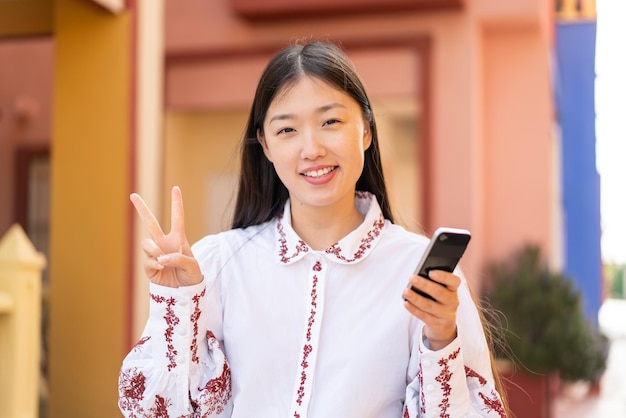 Junge Chinesin mit Handy im Freien lächelnd und Victory-Zeichen zeigend