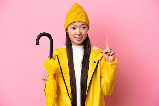 Junge Chinesin mit regenfestem Mantel und Regenschirm isoliert auf rosa Hintergrund, die einen Finger im Zeichen der Besten zeigt und hebt