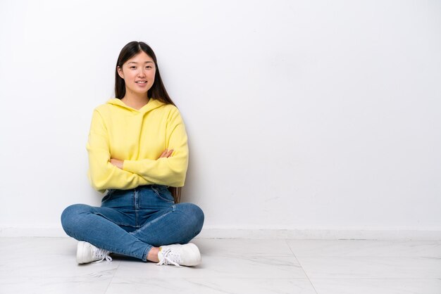 Junge Chinesin sitzt auf dem Boden isoliert an weißer Wand mit verschränkten Armen und freut sich