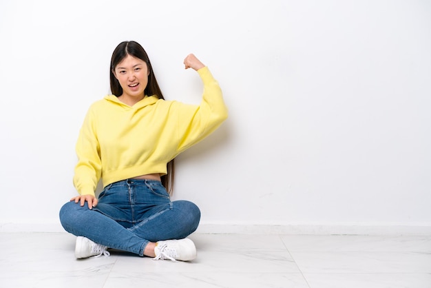 Junge Chinesin sitzt isoliert auf dem Boden an einer weißen Wand und macht eine starke Geste