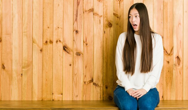 Junge chinesische Frau, die auf einem hölzernen Platz sitzt, der wegen etwas geschockt wird, das sie gesehen hat.