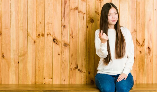 Junge chinesische Frau, die auf einem hölzernen Platz sitzt und mit dem Finger auf Sie zeigt, als ob die Einladung näher kommt.