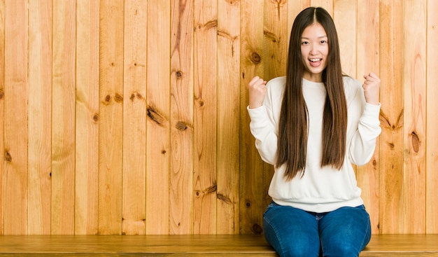 Junge chinesische Frau, die auf einem hölzernen Platz zujubelt sorglos und aufgeregt sitzt. Sieg-Konzept.