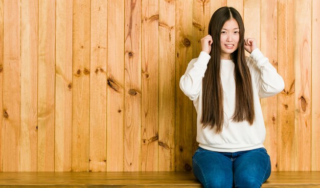 Junge chinesische Frau, die auf einem hölzernen Platzbedeckungsohren mit den Händen sitzt.