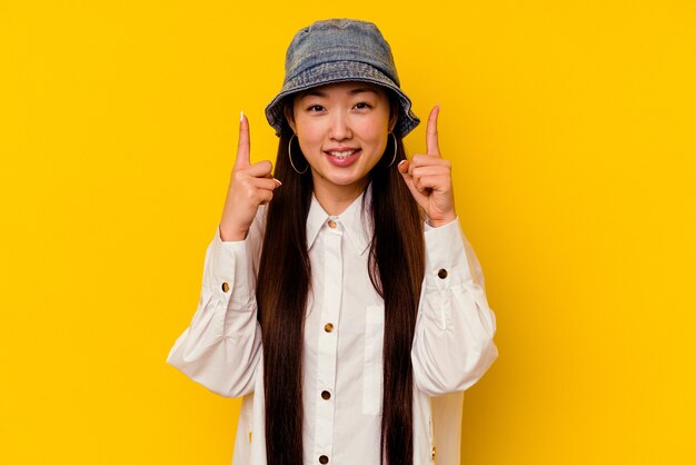 Junge chinesische Frau, die auf gelbem Hintergrund isoliert wird, zeigt mit beiden Vorderfingern an, die ein Leerzeichen zeigen.