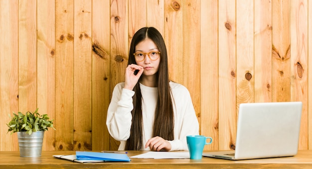 Junge chinesische Frau, die auf ihrem Schreibtisch mit den Fingern auf den Lippen ein Geheimnis halten studiert.