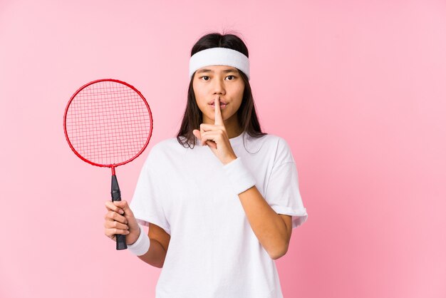 Junge chinesische Frau, die Badminton in einer rosa Wand hält ein Geheimnis oder bittet um Ruhe spielt.