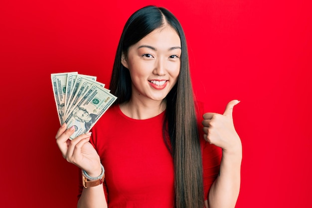 Junge chinesische Frau, die eine 20-Dollar-Banknote hält, lächelt glücklich und positiv, Daumen hoch und macht ausgezeichnetes und Genehmigungszeichen