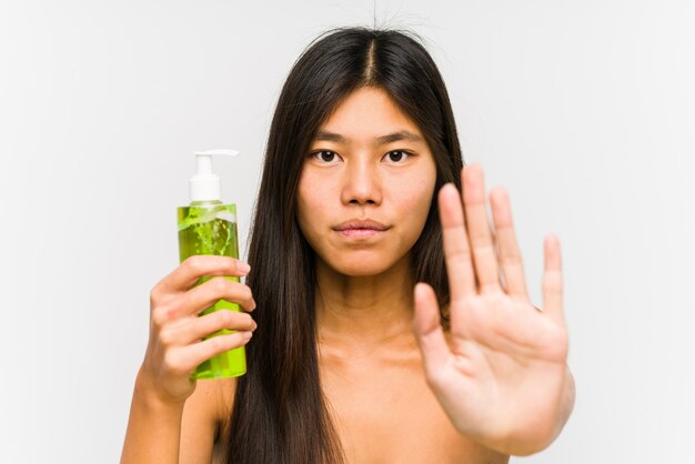 Junge chinesische Frau, die eine Feuchtigkeitscreme mit Aloe Vera hält, die mit ausgestreckter Hand steht, die Stoppschild zeigt, das Sie verhindert.