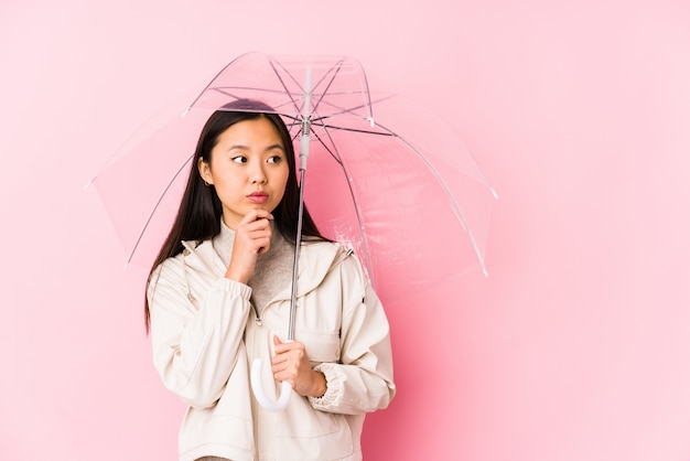 Junge chinesische Frau, die einen Regenschirm isoliert hält, der seitwärts mit zweifelhaftem und skeptischem Ausdruck schaut.