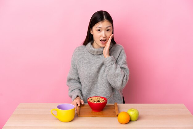 Junge chinesische Frau, die Frühstück in einem Tisch mit Überraschung und schockiertem Gesichtsausdruck hat