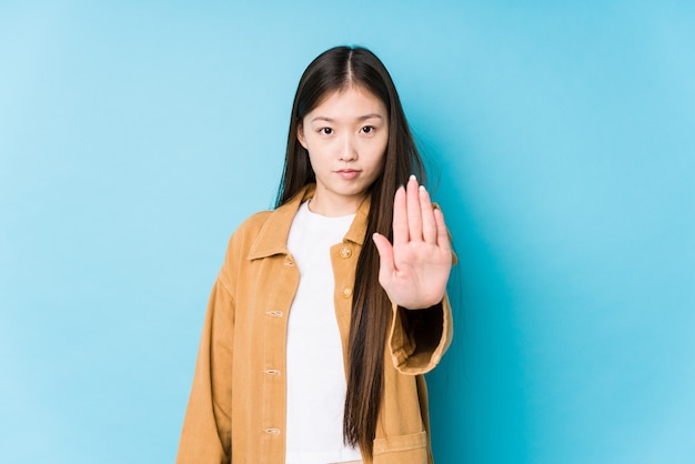 Junge chinesische Frau, die in einem blauen Hintergrund lokalisiert steht, der mit ausgestreckter Hand steht, die Stoppschild zeigt, das Sie verhindert.