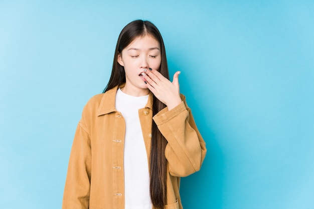 Junge chinesische Frau, die in einem isolierten Gähnen der blauen Wand aufwirft, zeigt eine müde Geste, die Mund mit Hand bedeckt.