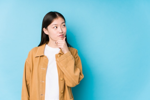 Junge chinesische Frau, die in einer blauen Wand isoliert seitlich mit zweifelhaftem und skeptischem Ausdruck aufwirft.